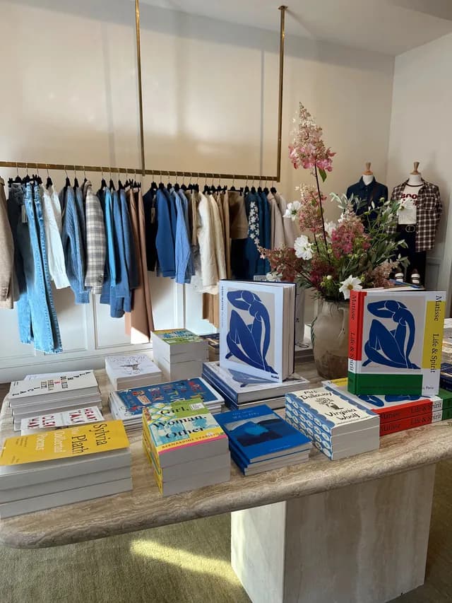Intérieur d’une boutique de proximité : portant de vêtements, table de livres et bouquet de fleurs